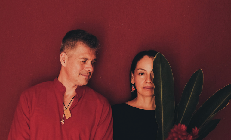Mexican acoustic guitar duo Rodrigo y Gabriela posing against a deep red wall with tropical plant leaves in a promo photo.